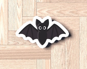 Può includere: Un adesivo a forma di pipistrello nero con occhi bianchi e un contorno bianco. L'adesivo è su uno sfondo di legno marrone chiaro.