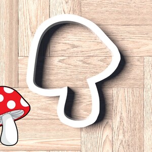May include: White plastic cookie cutter shaped like a mushroom. The cutter is on a wood background. A cartoon image of a red and white mushroom is in the lower left corner of the image.