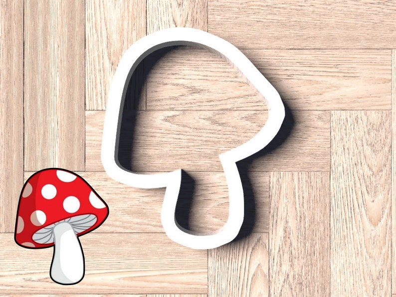 May include: White plastic cookie cutter shaped like a mushroom. The cutter is on a wood background. A cartoon image of a red and white mushroom is in the lower left corner of the image.