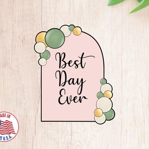 May include: A pink arch-shaped cookie cutter with the text "Best Day Ever" and a border of green and gold circles.