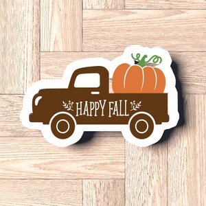 Pu&ograve; includere: Un adesivo a forma di camion marrone con le parole "HAPPY FALL" e una zucca nel cassone. La zucca &egrave; arancione con viticci verdi. L'adesivo &egrave; su una superficie di legno marrone chiaro.