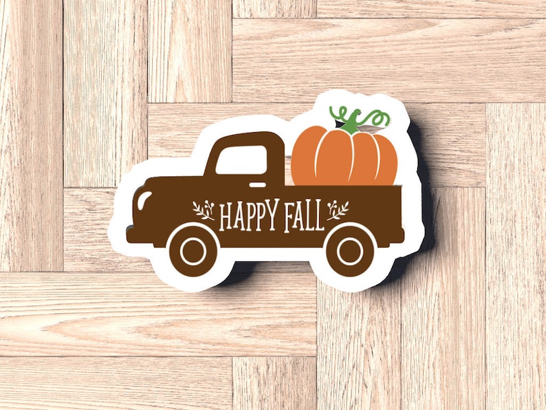 Pu&ograve; includere: Un adesivo a forma di camion marrone con le parole "HAPPY FALL" e una zucca nel cassone. La zucca &egrave; arancione con viticci verdi. L'adesivo &egrave; su una superficie di legno marrone chiaro.