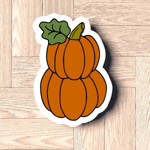 Puede incluir: Una pegatina de tres calabazas naranjas apiladas con una hoja verde en la parte superior. Las calabazas están delineadas en negro y tienen un fondo blanco.