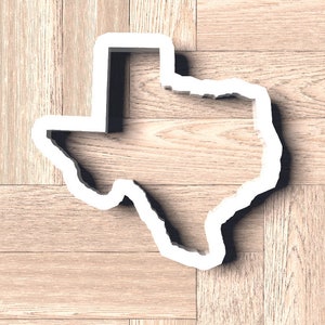 May include: A white outline of the state of Texas on a woodgrain background.