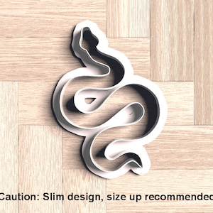 Può includere: Un tagliabiscotti bianco a forma di serpente. Il tagliabiscotti è realizzato in plastica e ha un design sottile. Il testo "Caution: Slim design, size up recommended" è stampato in basso nell'immagine.