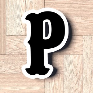 May include: Black letter P with a white outline, on a light wood background.