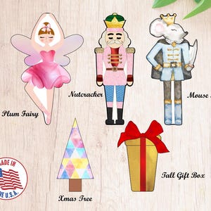 Könnte beinhalten: Ein Set mit fünf weihnachtlichen Ausstechern, darunter eine Zuckerpflaumenfee, ein Nussknacker, ein Mäusekönig, eine hohe Geschenkbox und ein Weihnachtsbaum. Die Ausstecher sind aus Metall und haben einen weißen Hintergrund mit bunten Designs.
