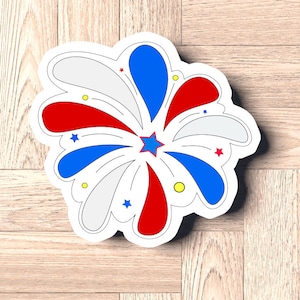 May include: A sticker featuring a stylized firework design. The design includes red, white, and blue elements, with star accents and a central starburst. The sticker is on a wooden surface.