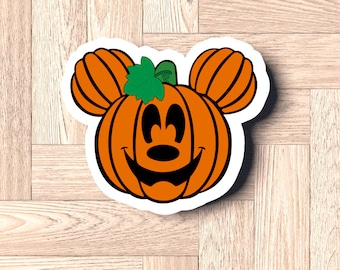 Può includere: Zucca arancione con un viso di Topolino, uno stelo verde e occhi neri. La zucca ha un contorno bianco ed è a forma di testa di Topolino.