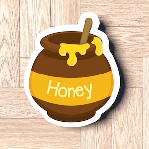 May include: A brown ceramic honey pot with a yellow lid and a wooden honey dipper. The pot is filled with honey and the word "Honey" is written on the front.