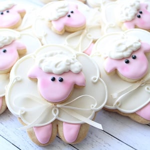 May include: A close-up of several pink and white sugar cookies decorated with white frosting and black eyes to look like sheep. The cookies are arranged on a white wooden surface.