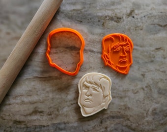 May include: Three cookie cutters, two orange and one white, shaped like human heads. The orange cutters are of a generic human head and a man with a serious expression. The white cutter is of a man with a serious expression.