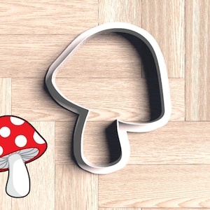 May include: White plastic cookie cutter shaped like a mushroom. The cutter is on a wooden surface. A red and white cartoon mushroom is in the lower left corner of the image.