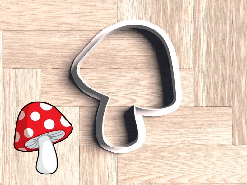 May include: White plastic cookie cutter shaped like a mushroom. The cutter is on a wooden surface. A red and white cartoon mushroom is in the lower left corner of the image.
