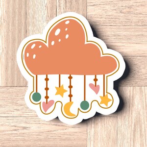 May include: A peach-colored cloud with a hanging mobile made of stars, hearts, and moons. The mobile is made of brown beads and has a green and peach color scheme.