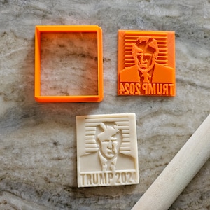 May include: An orange square cookie cutter and two cookie stamps. One stamp is orange and features a portrait with the text "TRUMP 2024". The other stamp is white and features the same portrait with the text "TRUMP 2024". A wooden rolling pin is also present.
