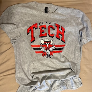 May include: A gray t-shirt with a red and black Texas Tech logo. The logo features the school mascot, a cartoon gunslinger, with the words "Texas Tech" above him.