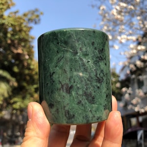 May include: A cylindrical, polished jade stone. The stone is a deep green color with darker green and black veining throughout. The object is held in a person's hand, with a blurred outdoor background.