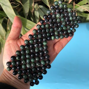 Op de afbeelding: Een verzameling donkergroene en zwarte ronde kralen, mogelijk jade, wordt getoond in de open hand van een persoon. De kralen zijn aan elkaar geregen, waardoor een textuur ontstaat. De achtergrond is een mix van groen gebladerte en een blauw oppervlak.
