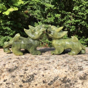 May include: Two jade green carved mythical creatures, possibly dragons or lions, face each other. The sculptures are detailed with textured surfaces and stand on a stone surface. The background features lush green foliage.