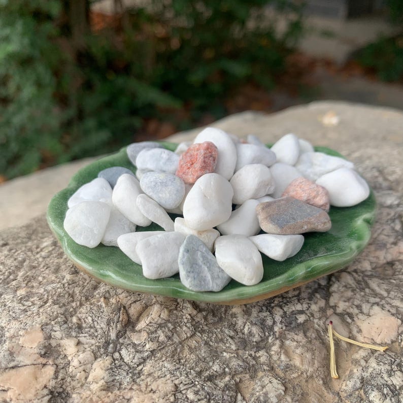 May include: A selection of smooth, polished stones in white, grey, and pink hues, presented on a green, leaf-shaped ceramic dish. The stones vary in size and texture, creating a natural aesthetic. The dish rests on a rough, textured stone surface.