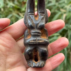 May include: A dark brown, carved figurine of a stylized figure with a rabbit-like head and a small hole at the base. The figurine has a weathered appearance with hints of orange-brown oxidation. The figure is held in a hand against a blurred green background.