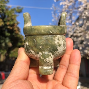 May include: A green jade censer with a weathered appearance. The vessel has two handles and is supported by three legs. Decorative patterns are carved into the sides of the censer. The background shows a blue sky and flowering tree.