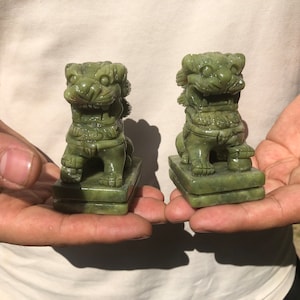 May include: Two jade green Foo Dog statues. Each statue is carved in detail, depicting a lion-like creature with a fierce expression, seated on a rectangular base. The statues are held in a person's hands.