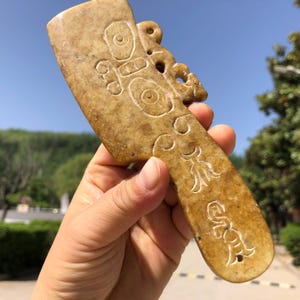May include: A tan-colored, ancient jade artifact with intricate carvings. The jade has a rectangular handle and a decorative top with circular and floral designs. The artifact is held against a backdrop of a blue sky and green foliage.