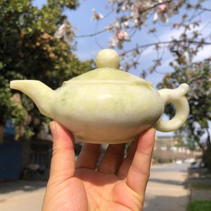 May include: A small, pale green stone teapot. The teapot features a rounded body, a curved spout, and a loop handle. A small, round knob is on the lid. The teapot is held in a hand, with trees and sky in the background.