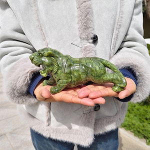Op de afbeelding: Een uit groene steen gesneden tijgerbeeldje met een gepolijste afwerking, voorzien van gedetailleerde gegraveerde strepen, een wijd opengesperde bek en een hurkende houding. Het beeldhouwwerk vertoont natuurlijke kleurvariaties in de steen.