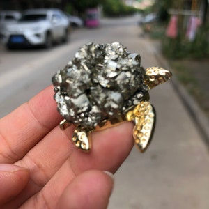 Puede incluir: Una figura de tortuga de pirita con base y patas de metal dorado. El caparazón de la tortuga está compuesto por muchos cristales pequeños, reflectantes y de aspecto metálico. Las patas y la cabeza son de color dorado brillante.