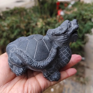 May include: A dark grey, carved stone turtle figurine. The turtle has detailed etchings on its shell and body, with its head raised and mouth open. The figurine is held in a person's hand, with a blurred green background.