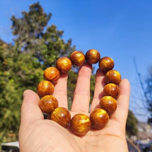 Puede incluir: Una pulsera de cuentas de color marrón dorado con una superficie pulida y reflectante. La pulsera está compuesta por cuentas esféricas, cada una de ellas con un patrón único de tonos claros y oscuros. La pulsera se sostiene en una mano sobre un fondo de cielo azul y follaje verde.