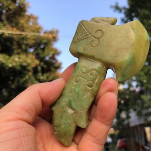 May include: A carved jade axe with a green and yellow hue. The handle features a fish-like design, and the axe head has intricate carvings. The axe is held in a hand, with a blurred background of trees and sky.