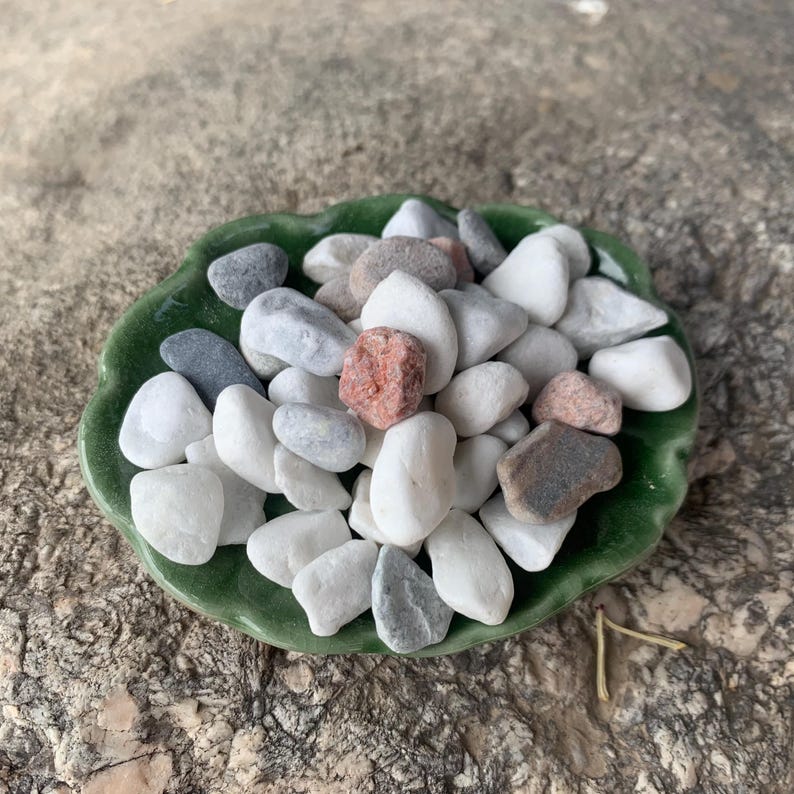 May include: A selection of smooth, polished stones in shades of white, grey, and reddish-brown, presented in a green, leaf-shaped ceramic dish. The stones are of varying sizes and shapes, offering a natural, organic appearance.