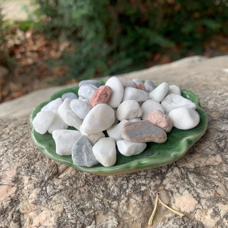 May include: A green, leaf-shaped ceramic dish filled with a variety of smooth, rounded stones. The stones are in shades of white, grey, and pink. The dish is placed on a textured stone surface, creating a natural display.
