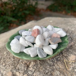 May include: A selection of smooth, polished stones in white, grey, and pink hues, presented in a small, green, leaf-shaped ceramic dish. The stones vary in size and shape, creating a natural, textured appearance.