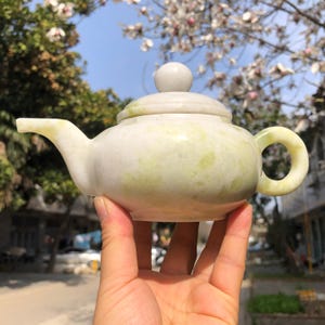 Op de afbeelding: Kleine, lichtgroene en witte stenen theepot met een afgeronde body, een gebogen tuit en een cirkelvormige handgreep. Het deksel heeft een kleine, ronde knop. De theepot wordt in een hand gehouden.