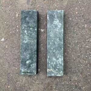 Puede incluir: Dos bloques de piedra rectangulares de color verde grisáceo con un patrón moteado. Los bloques están uno al lado del otro, mostrando una superficie natural y texturizada. La piedra tiene un tono verde oscuro con vetas verdes y blancas más claras.