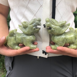 May include: Two jade-colored sculptures of mythical creatures, possibly dragons or lions, are held side-by-side. The carvings have detailed features, including manes, claws, and open mouths. The sculptures are approximately 5 inches long.