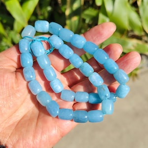 Könnte beinhalten: Eine Kette aus hellblauen Aquamarin-Edelsteinperlen. Die Perlen sind fassförmig und mit einer hellblauen Kordel aufgefädelt. Die Perlen werden in einer Hand vor einem Hintergrund aus grünem Laub gehalten.