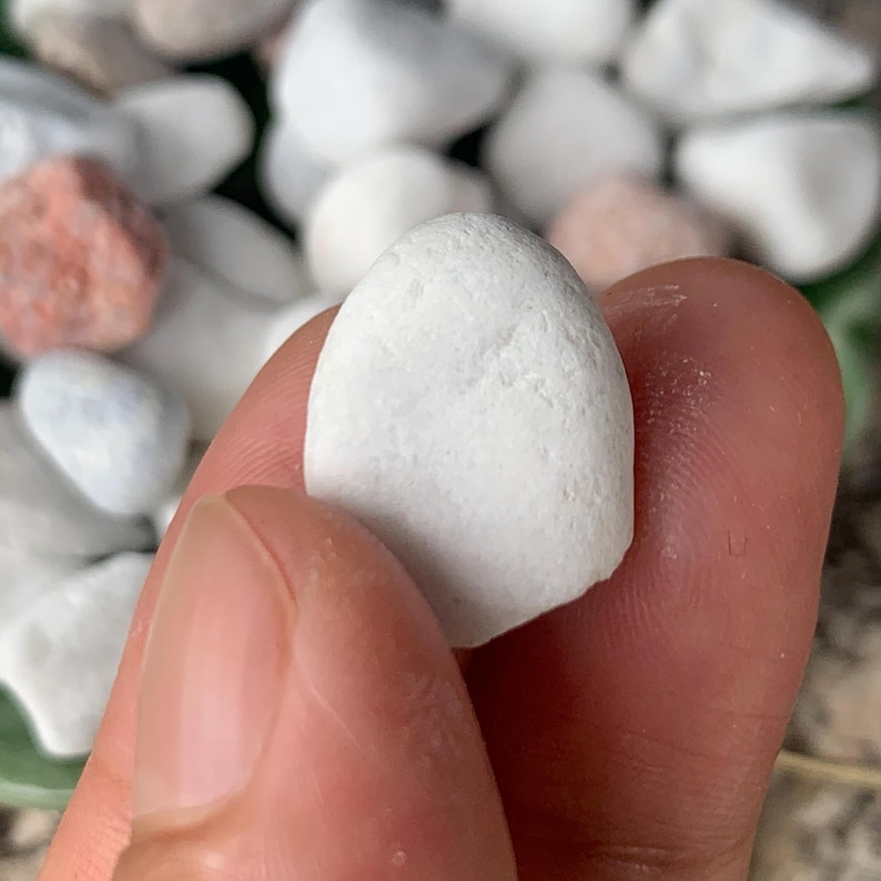 May include: A close-up of a smooth, white, oval-shaped stone held between fingers. The stone has a slightly textured surface. Other stones of various colours, including white and pink, are visible in the background.