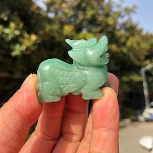 Puede incluir: Pequeña figura verde jade de una criatura mítica. La figura tiene un diseño detallado, con alas talladas y un cuerpo estilizado. El color es un verde suave y translúcido, sostenido en una mano.