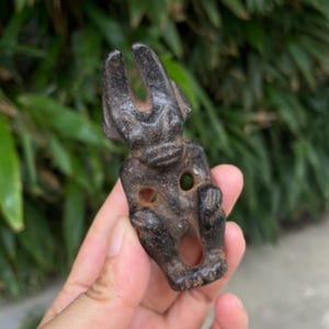Puede incluir: Una talla de piedra texturizada de color marrón oscuro que representa una figura antropomórfica estilizada con cuernos prominentes curvados hacia arriba, una boca ancha y recortes circulares en el pecho y la parte inferior.