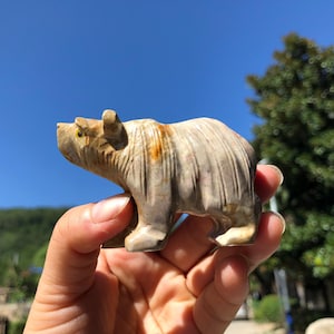 Puede incluir: Una figura de oso de piedra tallada en tonos beige y marrón. El oso se representa de pie, con una textura de pelaje detallada. La figura se sostiene en una mano sobre un fondo de cielo azul y follaje verde.