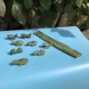 Op de afbeelding: Een verzameling kleine, gebeeldhouwde jade beeldjes, waaronder schildpadden, vissen en andere dieren, samen met verschillende lange, rechthoekige jade stukken. De beeldjes en jadestukken hebben een gevlekte groene kleur en zijn gerangschikt op een lichtblauwe ondergrond.