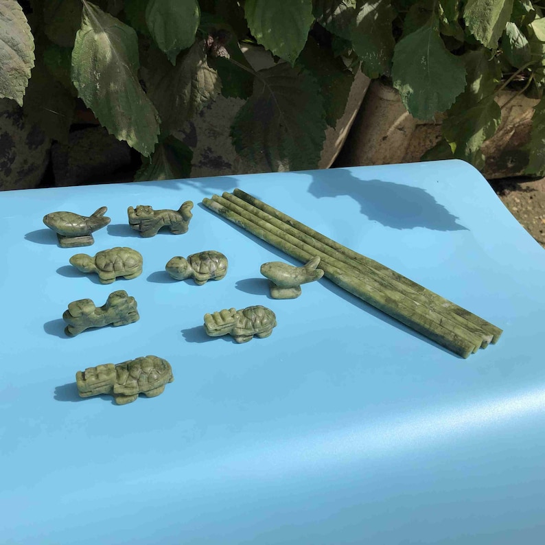 Op de afbeelding: Een verzameling kleine, gebeeldhouwde jade beeldjes, waaronder schildpadden, vissen en andere dieren, samen met verschillende lange, rechthoekige jade stukken. De beeldjes en jadestukken hebben een gevlekte groene kleur en zijn gerangschikt op een lichtblauwe ondergrond.
