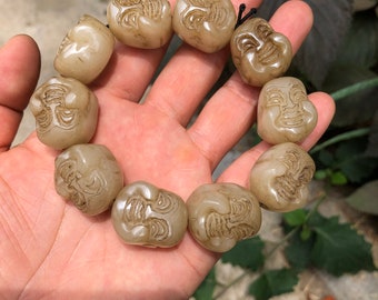 Op de afbeelding: Een armband van lichtbeige, gesneden kralen, elk met een lachend gezichtsontwerp. De kralen zijn geregen op een zwart koord. De armband wordt in een hand gehouden, waarbij de details van de gebeeldhouwde gezichten worden getoond.