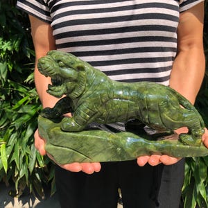 Könnte beinhalten: Eine grüne Jade-Tiger-Skulptur. Der Tiger befindet sich in einer brüllenden Pose, mit offenem Maul und sichtbaren Zähnen. Die Skulptur ist aus einem einzigen Stück Jade geschnitzt und ruht auf einem Sockel. Das Fell des Tigers ist mit geschnitzten Linien detailliert.